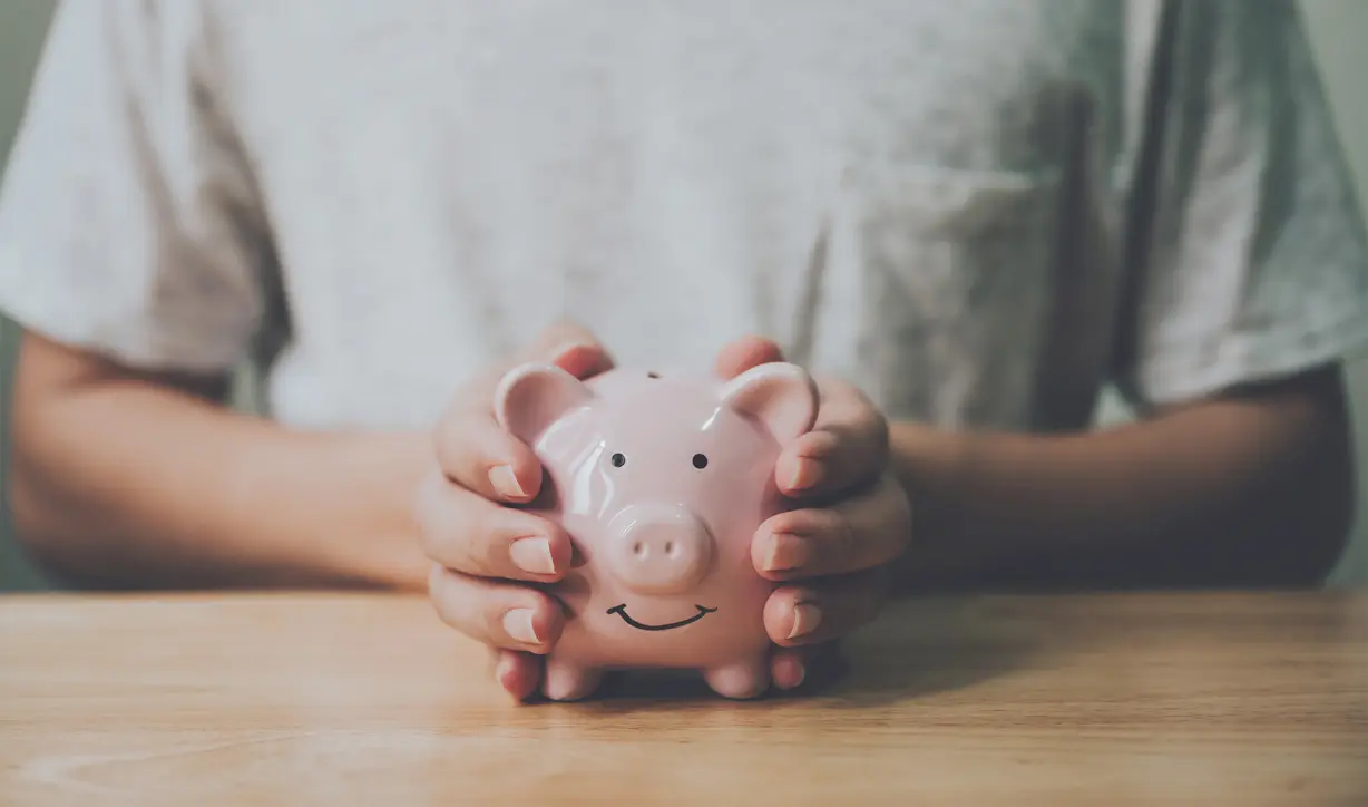 Budgetplanung Immobilie: Hände halten ein rosafarbenes Sparschwein über einem Holztisch