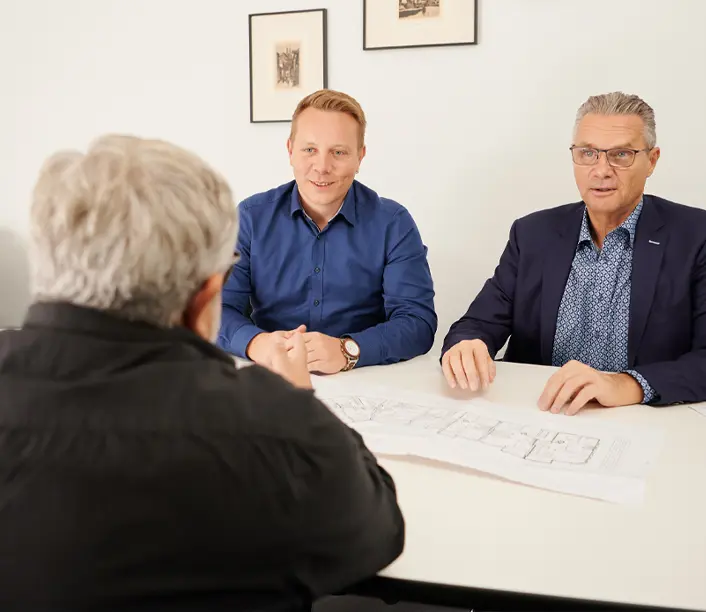 Zwei Berater sitzen einem Kunden gegenüber, auf dem Tisch liegen Pläne
